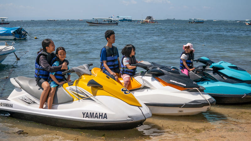 A family prepares to ride a jet ski on a Bali beach with a view of ships in the background.