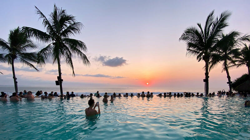 Tourists enjoy the sunset in an infinity pool with palm trees during high seasons Best Time To Visit Bali