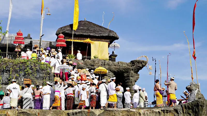 Balinese Hindus celebrating the Piodalan ceremony at Tanah Lot Temple, dressed in traditional attire and making offerings.