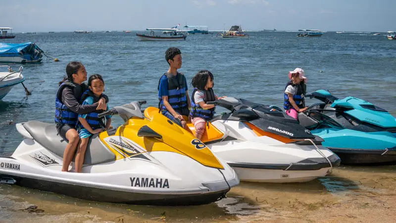 A family prepares to ride a jet ski on a Bali beach with a view of ships in the background