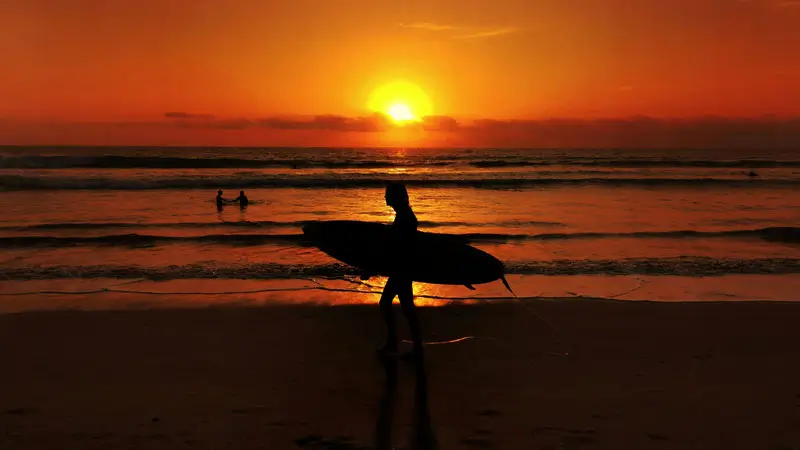 Surfers enjoy a stunning sunset at Kuta Beach, Bali with an orange sky. One of Natural Tourism Destinations in Bali