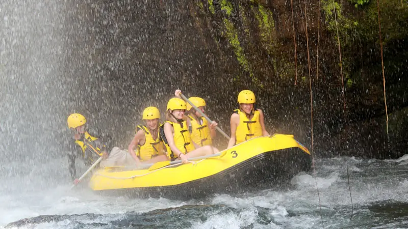 Rafting participants under a waterfall on Telaga Waja River in Bali, wearing yellow helmets and life vests.